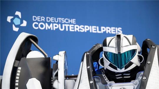 Der Gamescom Bot steht vor der Fotowand mit Schriftzug „Der Deutsche Computerspielpreis“.