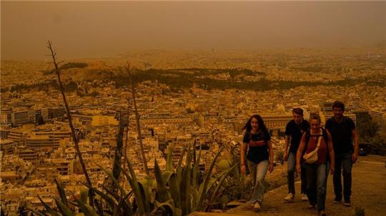 Touristen spazieren auf dem Lycabettus-Hügel, während der Himmel über Athen mit dem antiken Akropolis-Hügel im Hintergrund infolge von Saharastaub eingetrübt ist.
