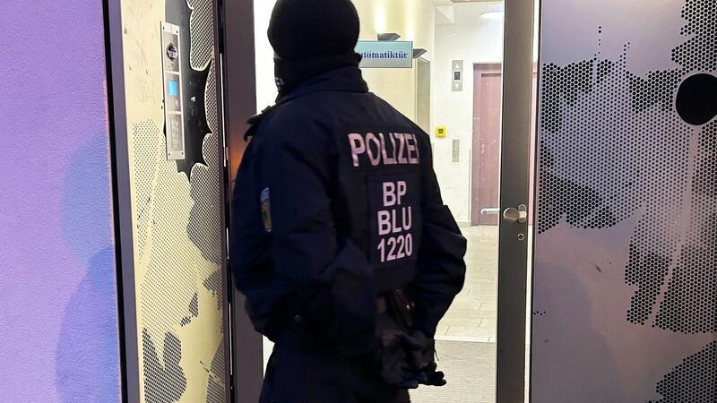 Ein Polizeibeamter steht bei einer Razzia in Berlin-Adlershof vor einem Gebäude.
