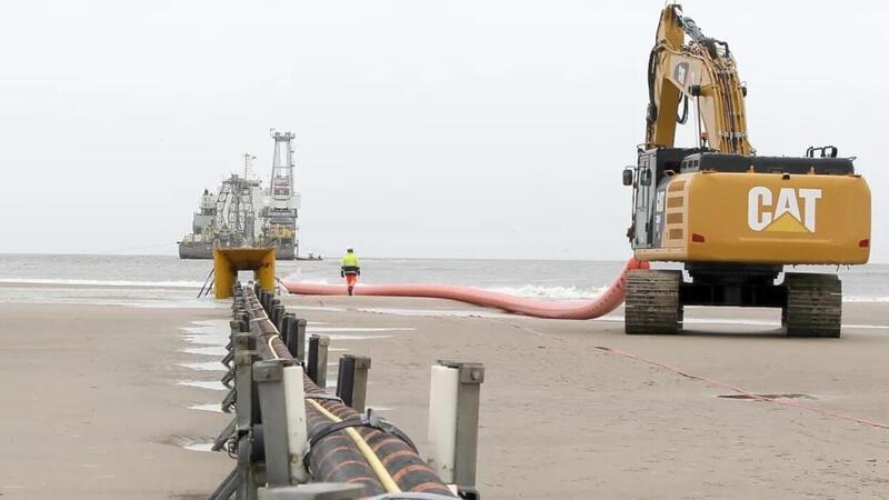 Anschläge und Cyber-Attacken – Die Offshore-Branche hat Angst Am Strand von Norderney verschwindet ein Stromkabel im Meer. Im Hintergrund ein Verlegeschiff, von dem aus der Kabelstrang auf den Meeresboden hinabgelassen wird, bis hinaus zu einem Windpark.