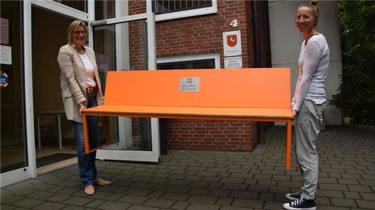 Amtsgerichtsdirektorin Nadine Romer-Moje (links) und Opferhelferin Denise de With tragen die orange Sitzbank in das Gerichtsgebäude. Das Möbelstück erhält seinen Platz vor dem Hauptsitzungssaal.