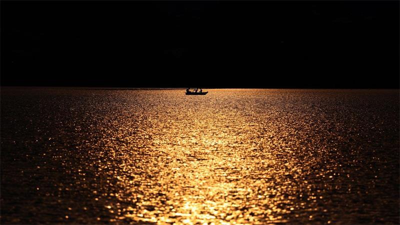 Angler rudern in einem Kahn über den im Sonnenuntergang glitzernden Hopfensee.