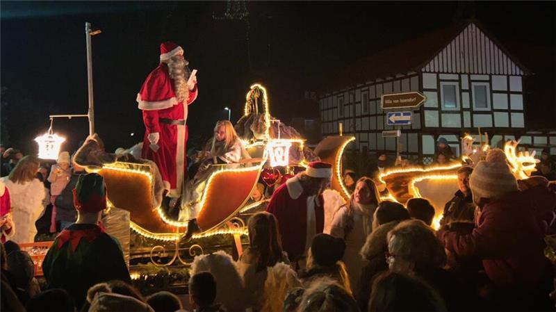 Der Weihnachtsmann steht aufrecht in einem mit Lichterschläuchen beleuchteten Schlitten. Davor steht eine Menschenmenge. 