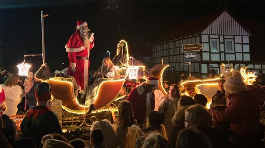 Der Weihnachtsmann steht aufrecht in einem mit Lichterschläuchen beleuchteten Schlitten. Davor steht eine Menschenmenge. 