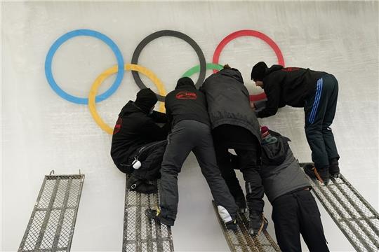 Arbeiter bringen im Vorfeld der Olympischen Winterspiele 2022 einen Ausschnitt der olympischen Ringe auf der Eisbahn des Yanqing National Sliding Center an. Foto: Jae C. Hong/AP/dpa