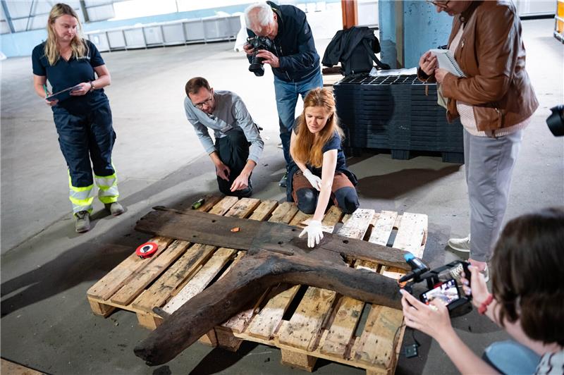 Bergung von Trave-Schiffswrack läuft gut – Erste Funde vorgestellt Archäologen erklären Journalisten die geborgenen Holzteile. In einer Lagerhalle wurden die ersten geborgenen Wrackteile eines Hanseschiffes präsentiert. Foto: Jonas Walzberg/dpa