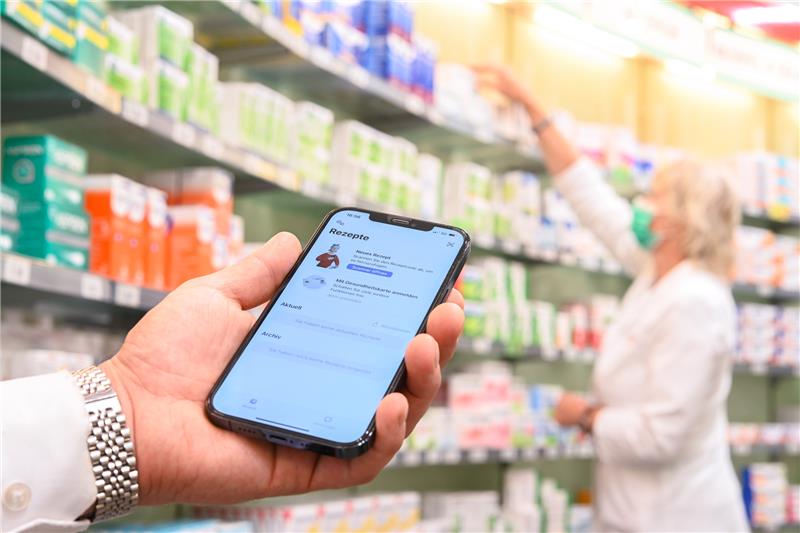 Auf einem Smartphone ist die geöffnete App «Das E-Rezept» zu sehen, während im Hintergrund eine Apothekerin in einer Apotheke an einem Regal steht. Foto: Mohssen Assanimoghaddam/dpa