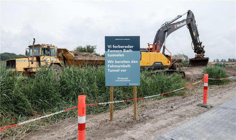 Baumaschinen heben hinter einem Schild mit der deutsch, dänischen Aufschrift «Wir bereiten den Fehmarnbelt-Tunnel vor» einen Entwässerungsgraben auf dem zukünftigen Baugelände aus. Foto: Markus Scholz/dpa