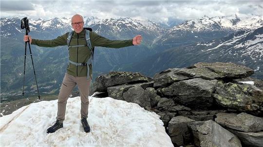 Oliver Strotmann steht mit ausgebreiteten Armen auf einer schneebedeckten Bergklippe mit dem Alpenpanorama im Hintergrund