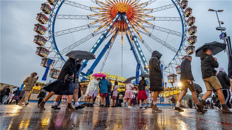 Besucher des Oktoberfestes in München gehen im Regen über das Festgelände vor dem Riesenrad.