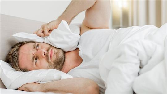 Ein Mann liegt im Bett und drückt sich ein Kopfkissen aufs Ohr.