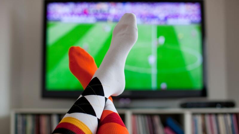 Immer mehr Fußballspiele: Schaut das noch wer? Überschlagene Beine vor einem TV-Gerät, auf dem ein Fußballspiel übertragen wird.