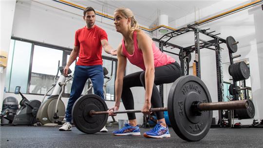 Eine Frau macht im Fitnessstudio Kreuzheben, ein Trainer hilft ihr.