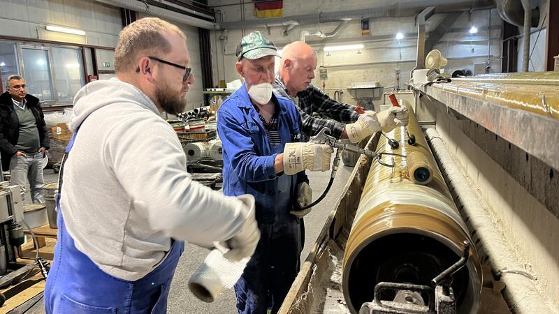 Manfred Bohm, Martín Monteserrín und Piotr Ochwat dicht hintereinander Schritt für Schritt enlang eines sich drehenden Kunststoffrohrs vor. Sie umwickeln es Runde um Runde mit Glasfaser-Gewebe, das mit Harz begossen wird.