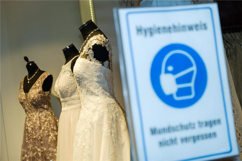 Brautkleider sind in einem Schaufenster eines Brautmoden-Geschäfts in der Innenstadt zu sehen. Im Vordergrund steht ein Schild, das auf die Maskenpflicht während der Corona-Pandemie verweist. Foto: Ole Spata/dpa