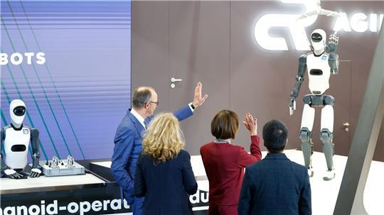 Bundeskanzler Friedrich Merz winkt einem Roboter zu – die Zukunft winkt zurück.