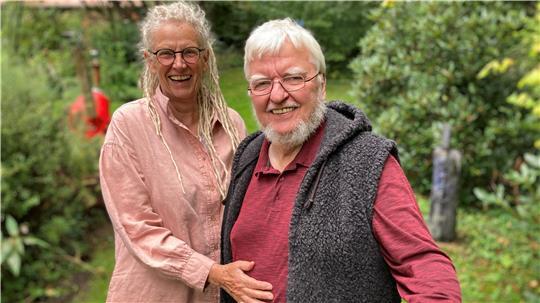 Das Künstlerpaar Christa Donatius und Michael Jalowczarz  steht mit einladener Geste in der grünen oase von Gut Daudieck