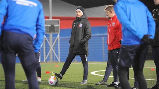Der Fußballtrainer der SV Drochtersen/Assel, Oliver Ioannou, steht neben seinen Spielern auf dem Rasen und spricht. Er hat einen Ball am Fuß.