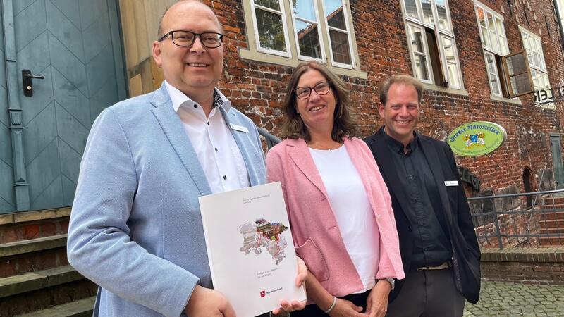 Das Mäppchen wiegt leicht, ist aber rund zwei Millionen Euro schwer: Stades Bürgermeister Sönke Hartlef hält den Förderbescheid in seinen Händen, den Karin Beckmann gerade an ihn und Planer Jens Bossen (rechts) übergeben hat.