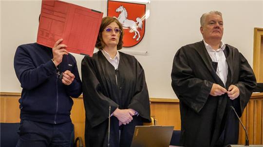 Der Angeklagte steht mit einem Aktenordner vor dem Gesicht neben seinen Verteidigern Dinah Busse und Dirk Meinicke im Saal des Landgerichts Stade.