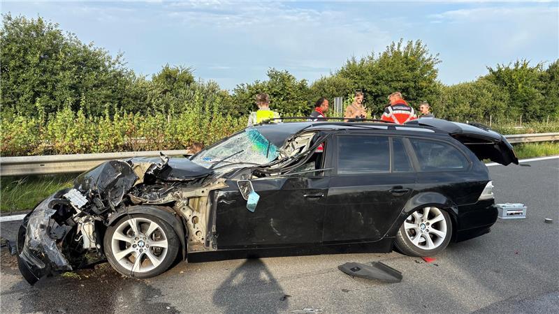 Seiten-Ansicht eines verunglückten BMWs auf der Autobahn A26 am Morgen des 12.8.2025. Hinter dem Fahrzeug stehen Unfall-Beteiligte, Zeugen und Mitarbeitende des Rettungsdienstes. 