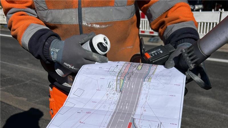 Der Blick auf den Plan, ein Zollstock, Farbspray und eine Schnurrolle sorgen dafür, dass die Fahrbahnmarkierungen an der richtigen Stelle auf den Asphalt kommen.