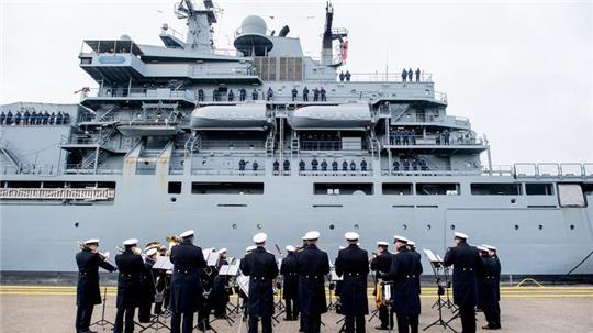 Der Marinemusikkorps Wilhelmshaven steht an einer Kaimauer, während die „Frankfurt am Main“ in den Hafen einfährt.