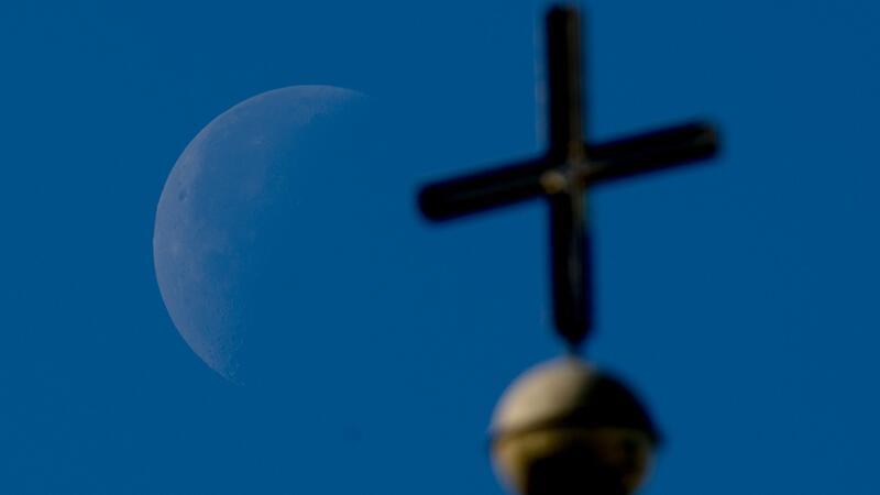 Der abnehmende Viertelmond ist hinter einem Kirchenkreuz zu sehen.
