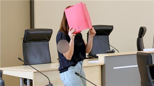 Die Angeklagte betritt den Gerichtssaal mit rotem Aktendeckel vor dem Gesicht.