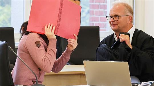 Die Angeklagte betritt den Gerichtssaal seit Monaten mit Fußfesseln und einem Aktendeckel vor dem Gesicht.