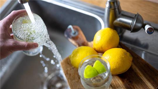 Trinkwasser läuft aus dem Wasserhahn in einer Küche in ein Glas mit Limettenstücken, während im Vordergrund Zitronen auf einem Küchenbrett liegen.
