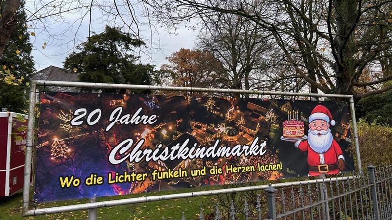 Ein Banner mit der Aufschrift "20 Jahre Christkindmarkt - Wo die Lichter funkeln und die Herzen lachen" steht im Park der Villa von Issendorff in Himmelpforten. Das Banner zeigt ein Luftbild des Christkindmarktes und einen freundlichen Comic-Weihnachtsmann mit einer Geburtstagstorte.