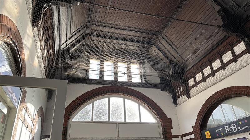 Die Decke der Bahnhofshalle ist mit dunklem Holz vertäfelt. Auf dem gespannten Netz gegen Tauben glitzert Staub in der Sonne.