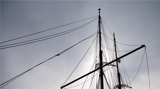 Die Masten eines Segelschiffes zeichnen sich bei trübem Wetter vor dem bewölktem Himmel ab.