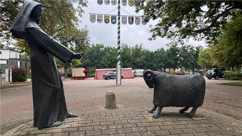 Die Nonne Mette zieht das Schaf Oskar: An der Skulptur auf dem Schafmarktplatz in Altkloster spenden Bäume Schatten. Im Hintergrund sind Verkaufsstände der Wochenmarktbeschicker zu sehen.