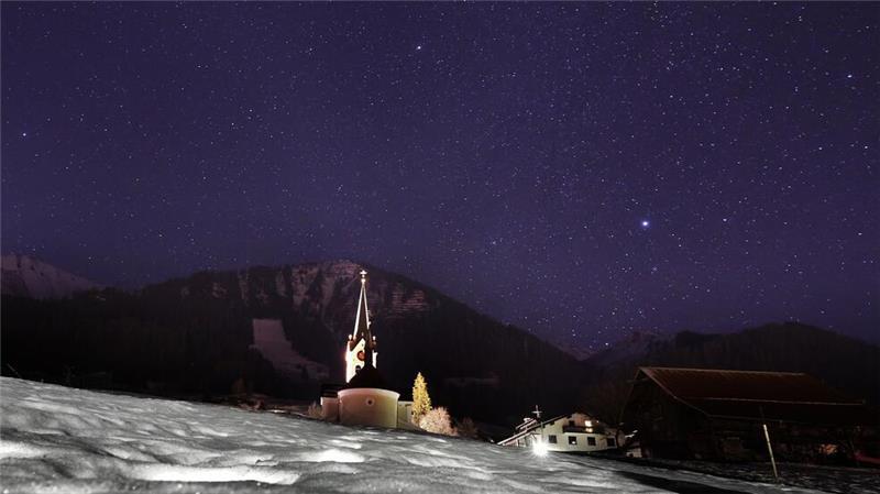 Die Pfarrkirche Mariä Opferung steht in Riezlern unter dem Sternenhimmel hinter einer schneebedeckten Wiese.