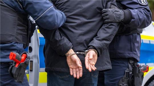 Zwei Polizisten stehen mit einem Mann in Handschellen vor einem Peterwagen.