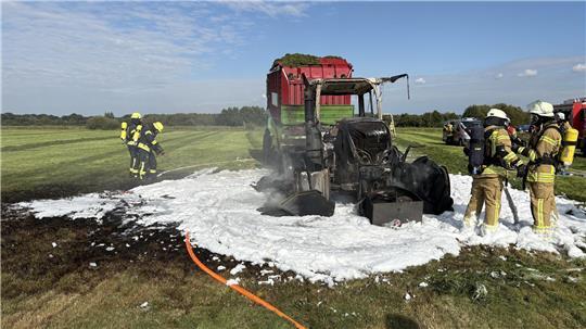 Einsatzkräfte der Feuerwehr neben einem ausgebranntem Traktor mit Anhänger auf einem Feld in Düring, Gemeinde Loxstedt. 
