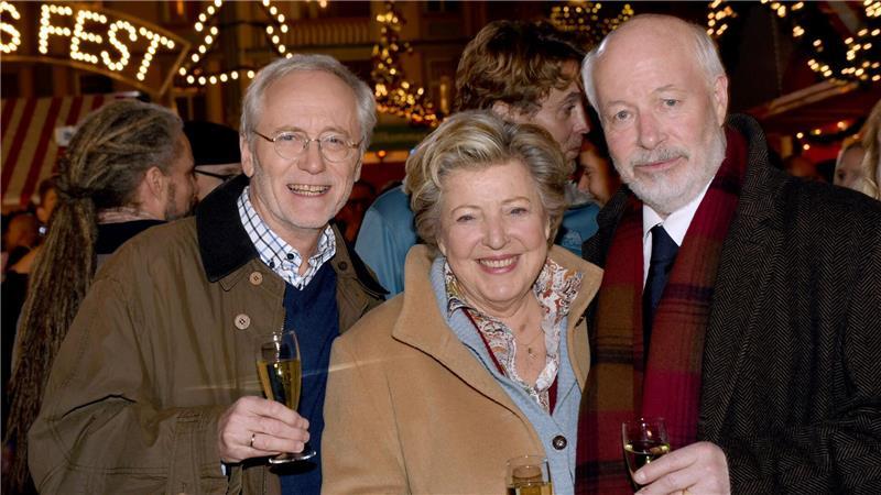 Die Schauspieler Joachim Hermann Luger (Hans Beimer, links) und die Schauspieler Marie-Luise Marjan (Mutter Beimer) und Bill Mockridge (Erich Schiller, rechts) posieren in der Außenkulisse mit Sektgläsern nach der Live-Ausstrahlung der 1559. Folge im Dezember 2015.