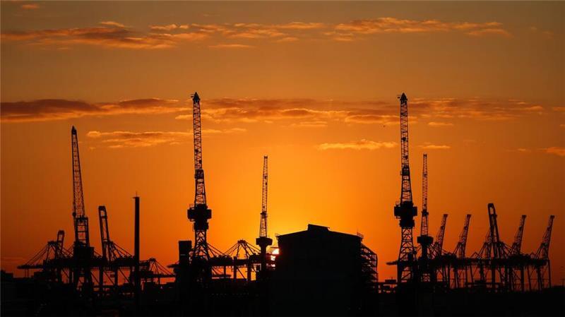 Die Sonne geht hinter Containerbrücken und Hafenkränen im Hamburger Hafen unter.