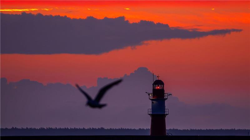 Die aufgehende Sonne färbt den Himmel hinter dem Leuchtfeuer auf der Ostmole am Ostseestrand von Warnemünde orangerot.
