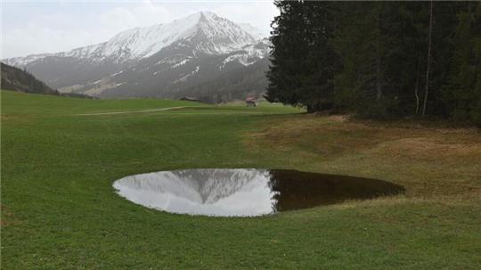 Die im Dunst liegenden Alpen spiegeln sich in einem Weiher. Der Saharastaub-Höhepunkt ist überschritten.