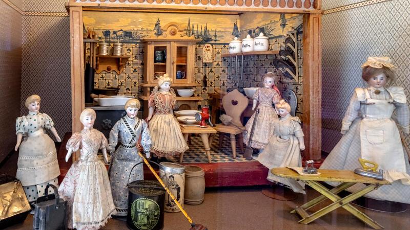 Von Porzellanpuppe bis Barbie: Auf Zeitreise im Puppenmuseum Falkenstein Eine Puppenstube, die eine Küche darstellt, mit sieben verschiedenen Puppen in historischen Kleidern.
