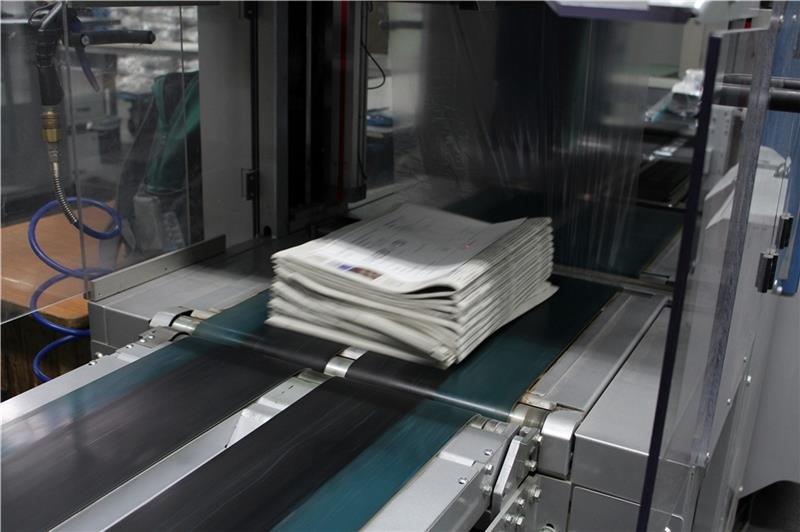Die paketweise gestapelten Zeitungen werden von Maschinen in Folie verpackt und mit Bändern zusammengebunden, um dann zu den Zustellern gebracht zu werden.