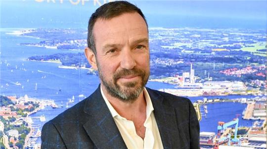 Dirk Claus, Geschäftsführer der Seehafen Kiel GmbH, steht vor einem Foto des Hafens.