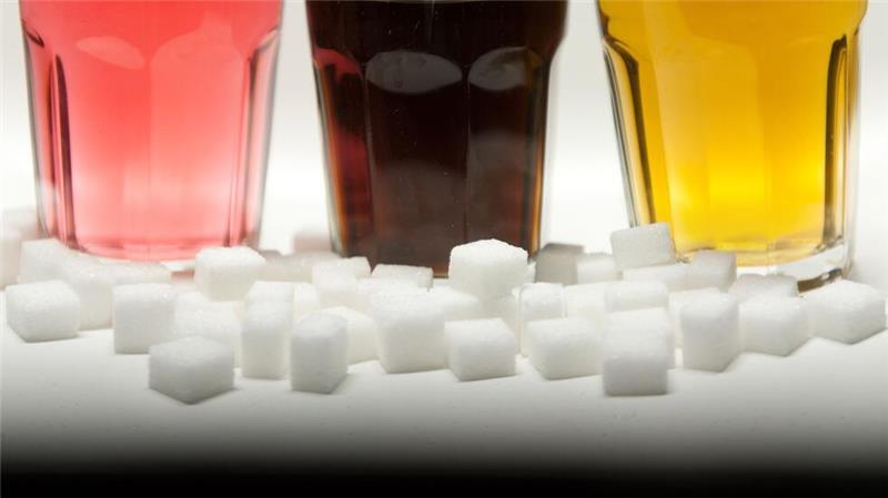 Drei Gläser gefüllt mit roter Limonade, Cola und einem Energy-Drink neben Zuckerwürfeln auf einem Tisch