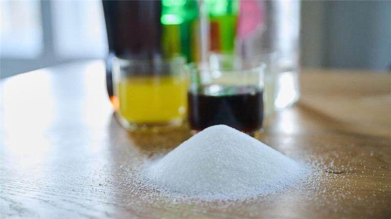 Drei unterschiedliche Arten Softdrinks und die Flaschen dazu stehen auf einem Tisch, während ein Berg von Zucker davor aufgeschüttet ist