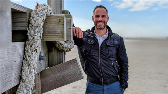 Blauer Himmel, schwarze Lederjacke: Daniele Palu lehnt an einem Pfahlbau am Strand von St.-Peter-Ording