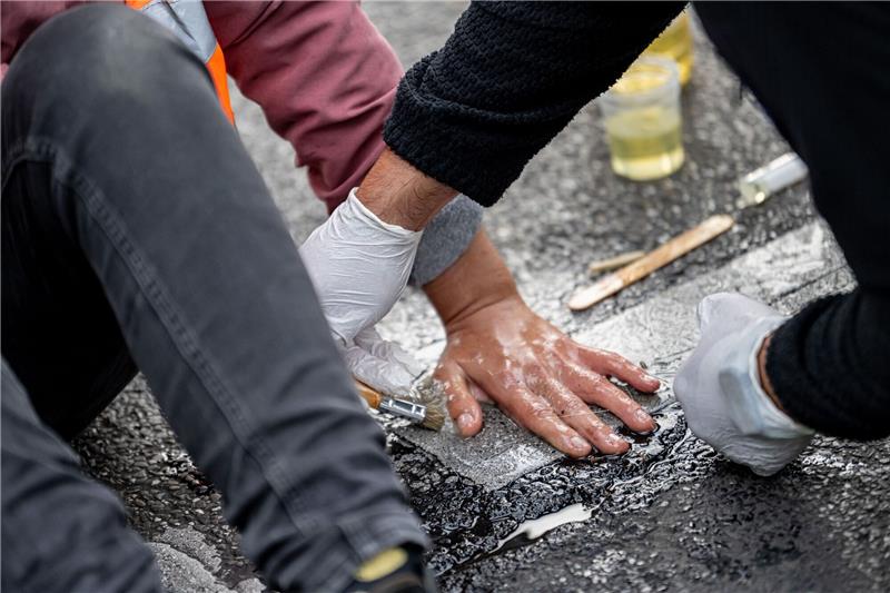 Eien Aktivistin der Letzten Generation hat sich auf die Straße geklebt. Ein Polizist versucht, ihre Hand vom Asphalt zu lösen.