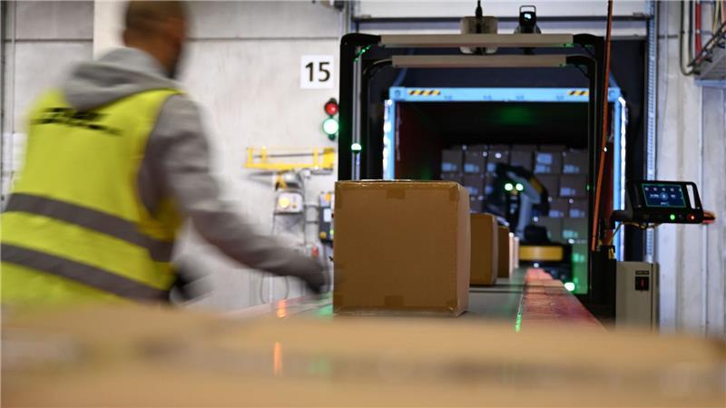 Ein DHL-Mitarbeiter nimmt Pakete in Emfpang - im Hintergrund ist ein Roboter zu sehen, der die Pakete aus einem Container nimmt und sie auf ein Förderband legt. (Archivbild)